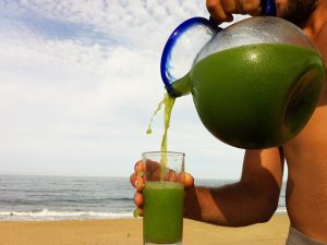 Close up of shirtless man pouring a glass of healthy green colored juice from a pitcher on the beach with the sand, sky and ocean