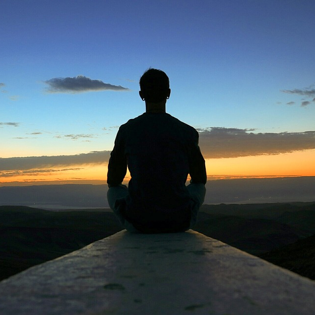 Young man meditation as he looks at a sunset, sitting on the end of a blockk wall with his back to the viewer.